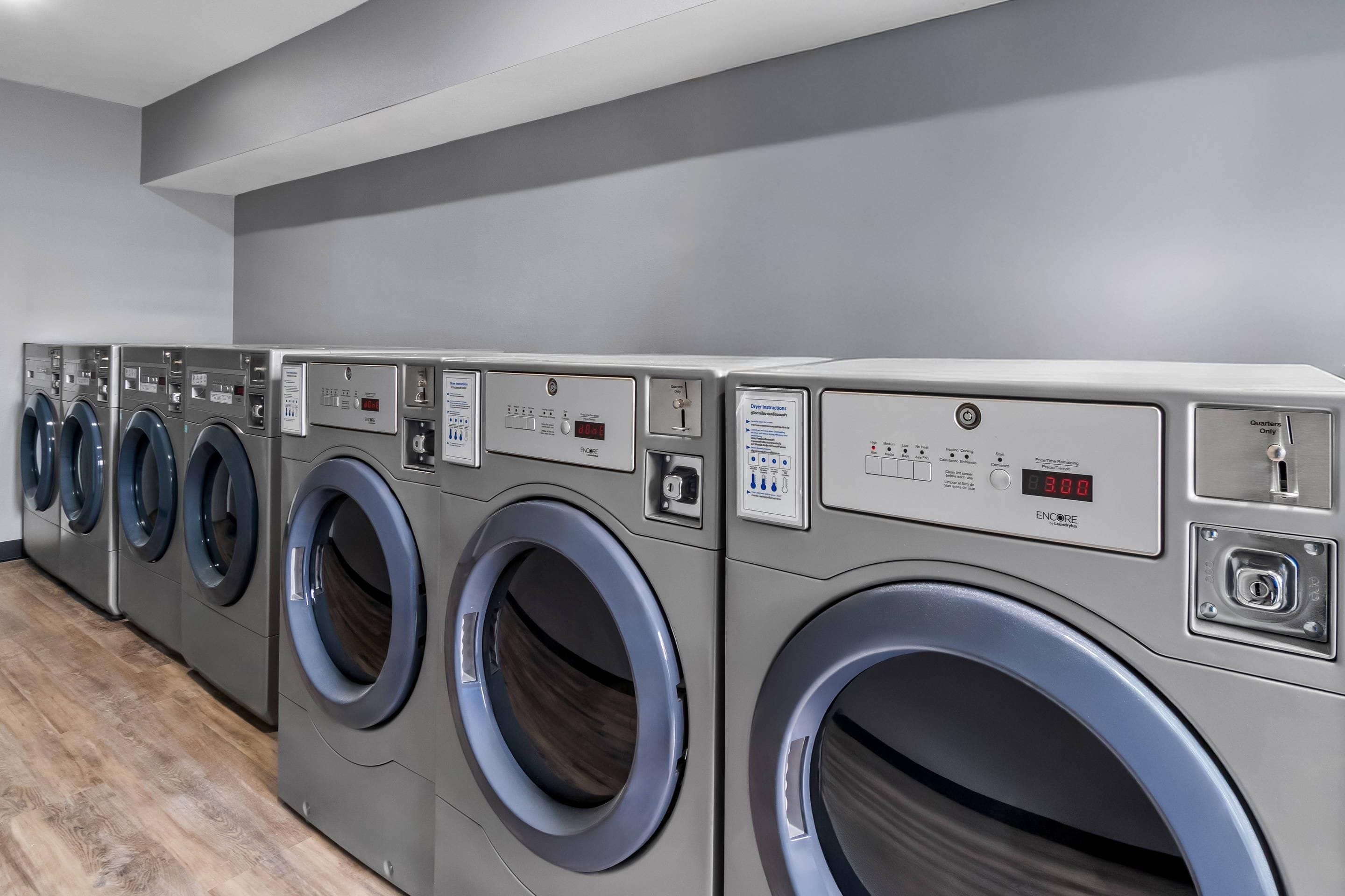 A row of front-loading washing machines in a modern laundry room with grey walls and wooden flooring.