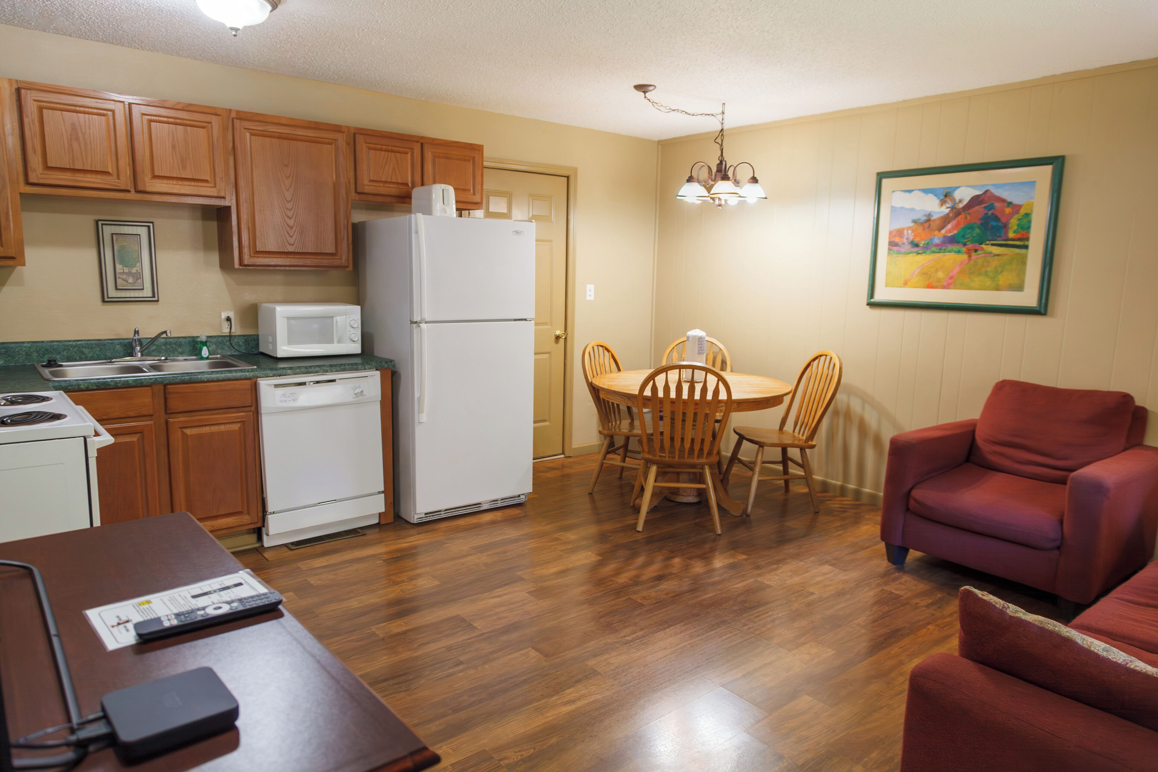 A compact kitchen with wooden cabinets, a white refrigerator, and a microwave. There is a dining table with four chairs and a red armchair. A painting is on the wall, and a remote control is on the table.