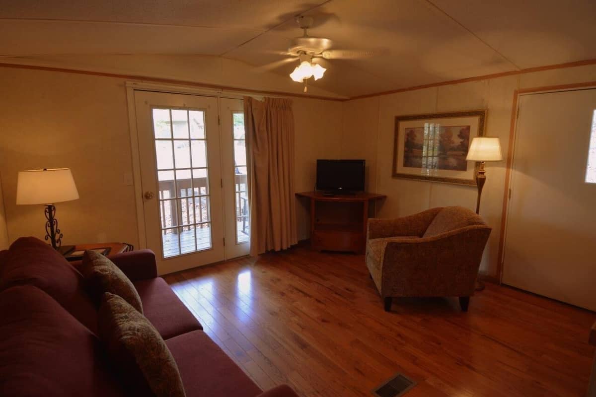 A living room with a wooden floor, a sofa, a chair, a television, a ceiling fan, and a door with a view.