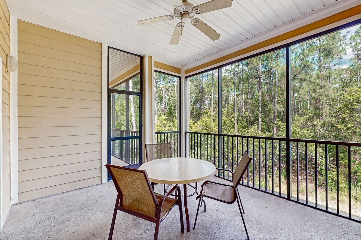 A covered outdoor area with a round table and chairs, large windows offering a view of a forest, and a ceiling fan.