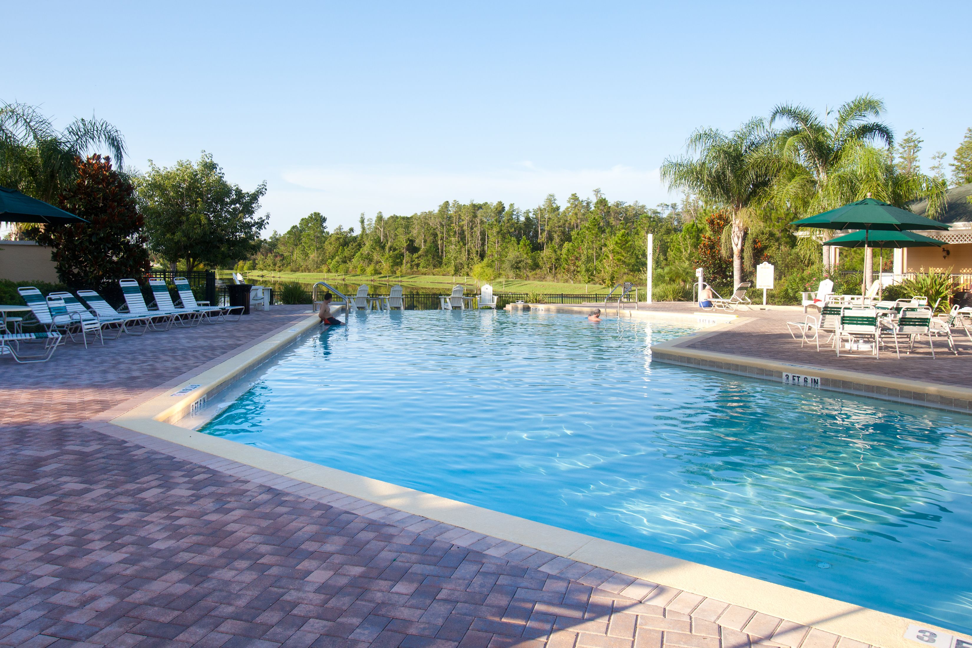 Una piscina rectangular con borde de ladrillo, rodeada de sillas de descanso y sombrillas, con un área verde y árboles al fondo.