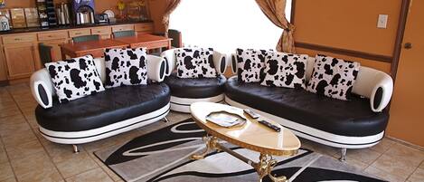 A living room with a black and white cow print sofa set, a coffee table, and a kitchen area in the background.
