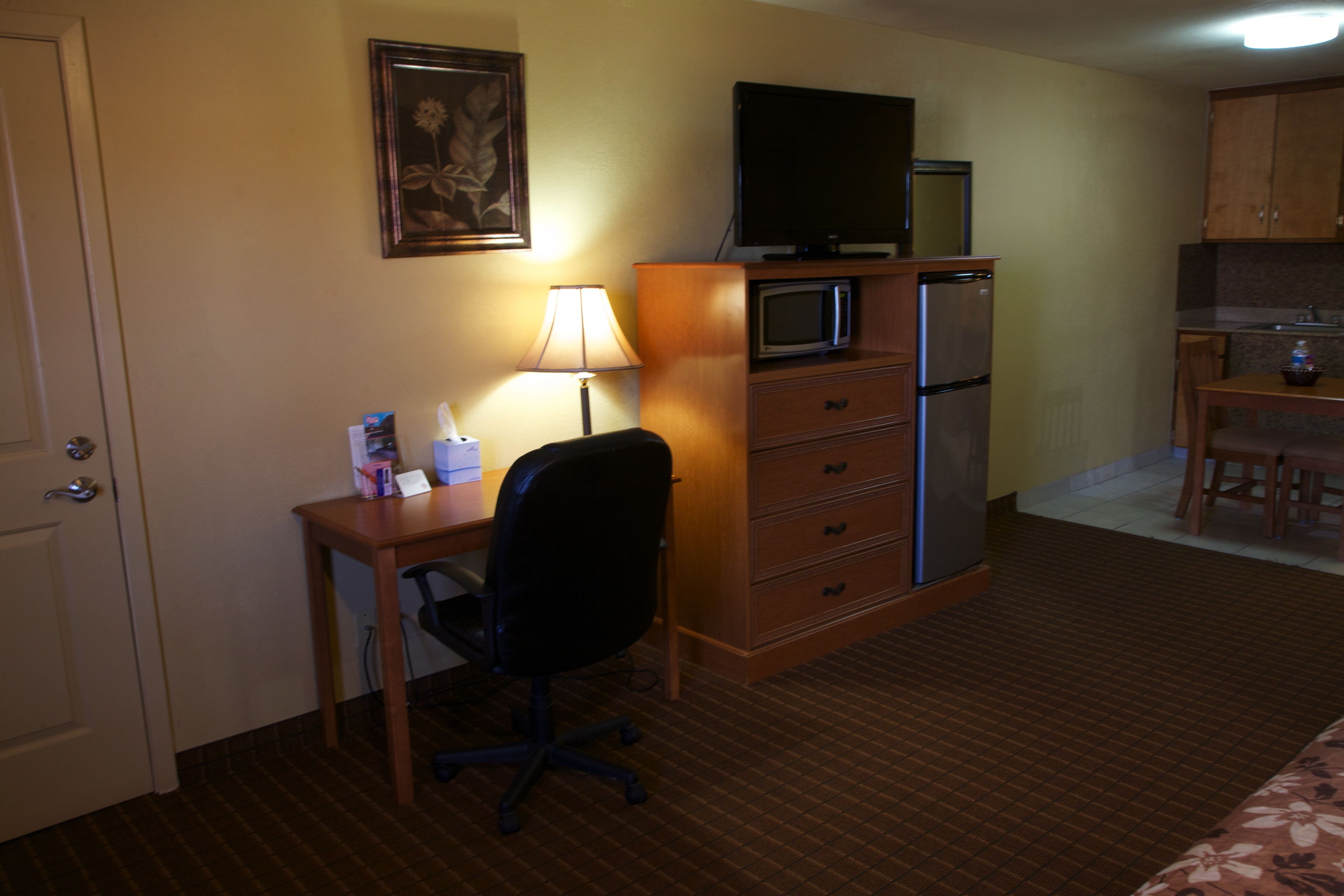 Habitación de hotel con escritorio, silla, lámpara, microondas, refrigerador y televisor sobre un mueble de madera.