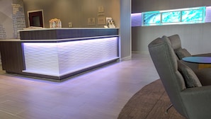 Ein Hotel-Empfangsbereich mit moderner Rezeption, einer Sitzecke mit rundem Tisch und einem wandmontierten digitalen Bildschirm.
