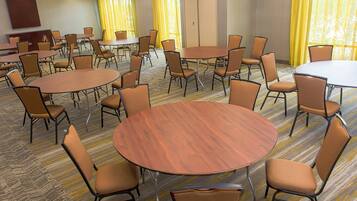 Un salón de conferencias con varias mesas redondas y sillas, suelo alfombrado y ventanas amplias con cortinas amarillas.