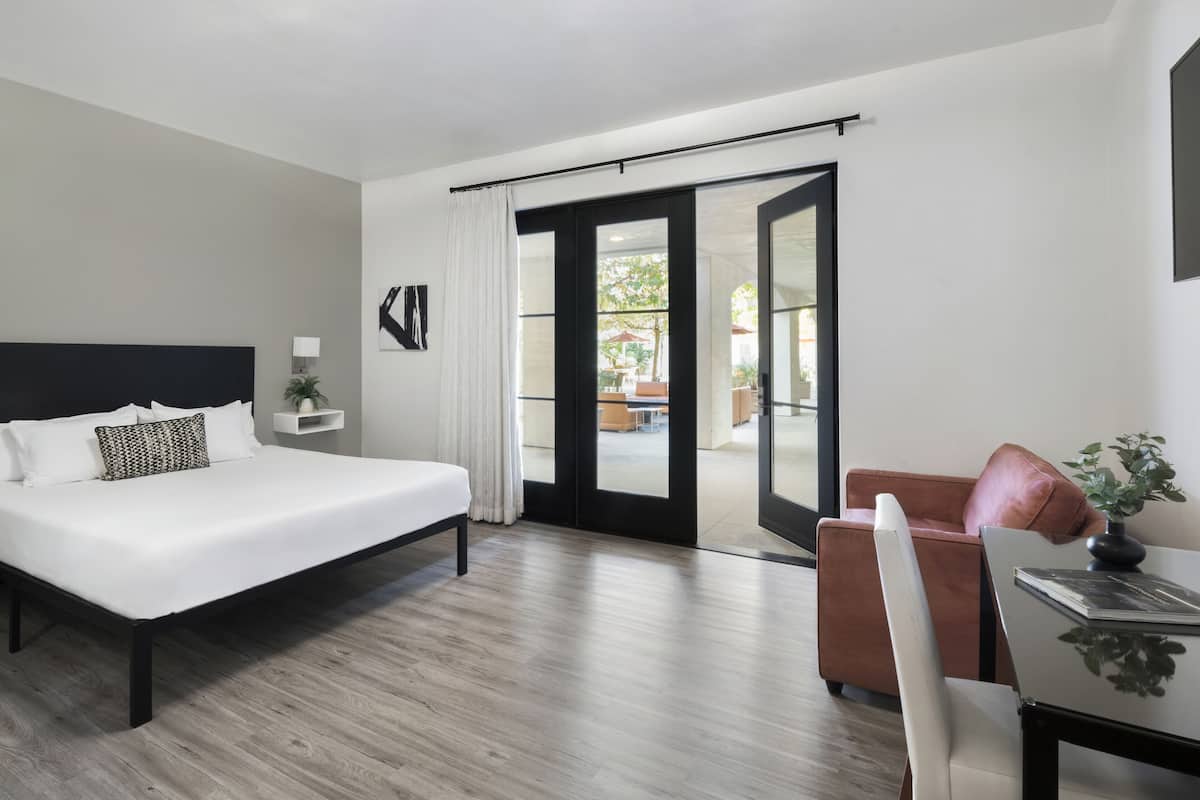 Una habitación de hotel moderna con una cama grande, un escritorio, una silla y un ventanal corredera que da a un espacio exterior.