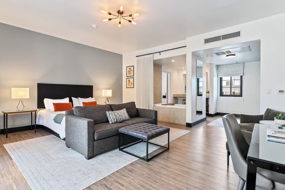 Una habitación de hotel moderna con un sofá gris, un otoman negro, una cama blanca y una mesita de noche con una lámpara.