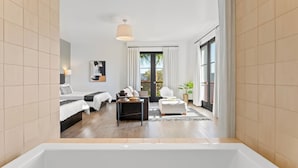 A modern hotel room with a large bathtub, a sofa, a coffee table, and a view of the outdoors through a large window.