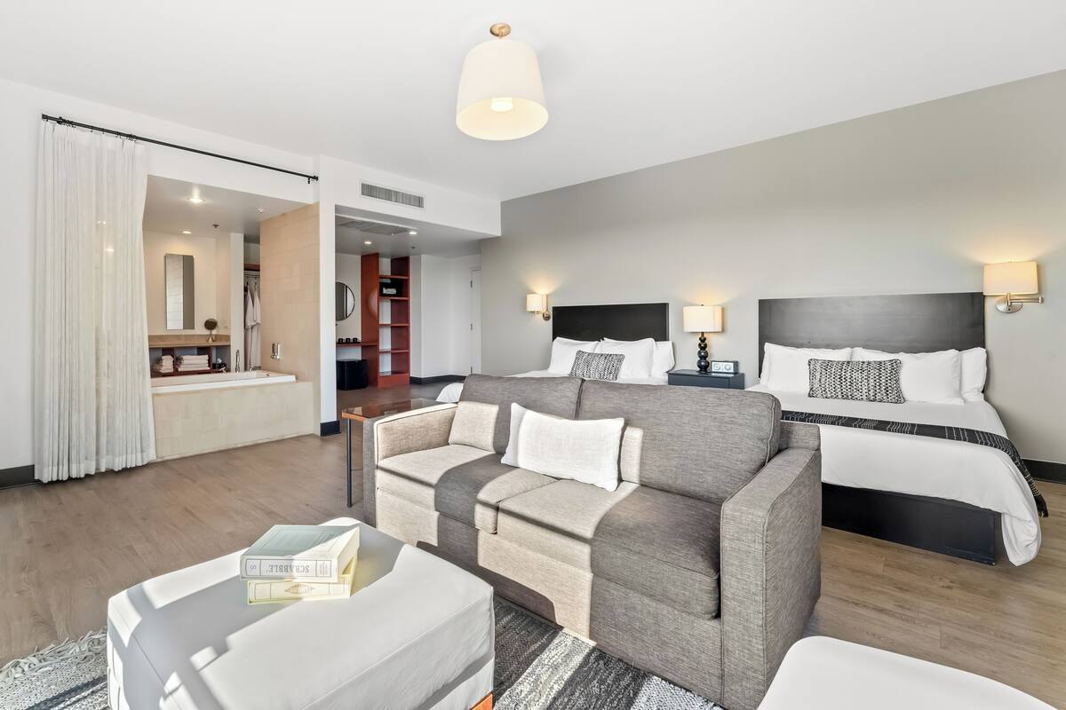 Una habitación de hotel moderna con un sofá gris, una cama con cabecera negra y una pequeña mesa con libros.