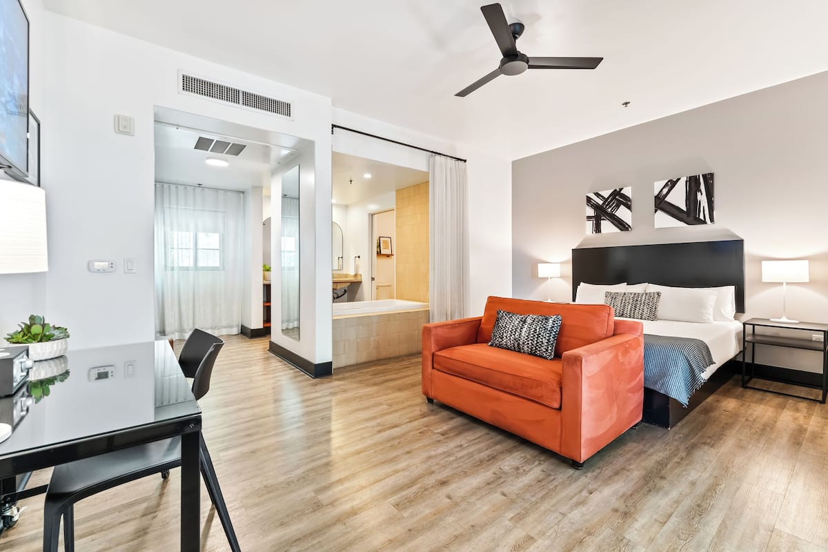 Una habitación de hotel moderna con piso de madera, una cama con ropa blanca, un sofá naranja, un escritorio con una silla y un ventilador de techo.