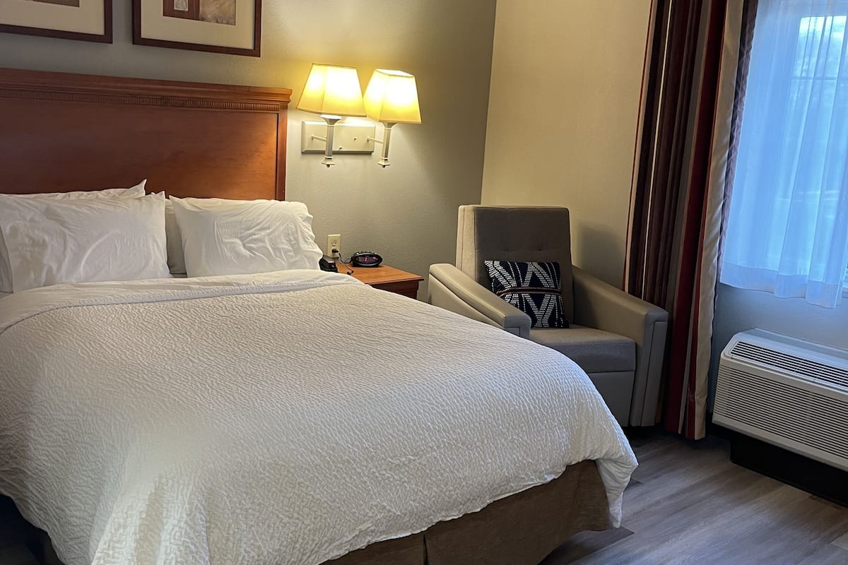 A hotel room with a large bed, two wall-mounted lamps, a chair, a radiator, and two framed pictures on the wall.