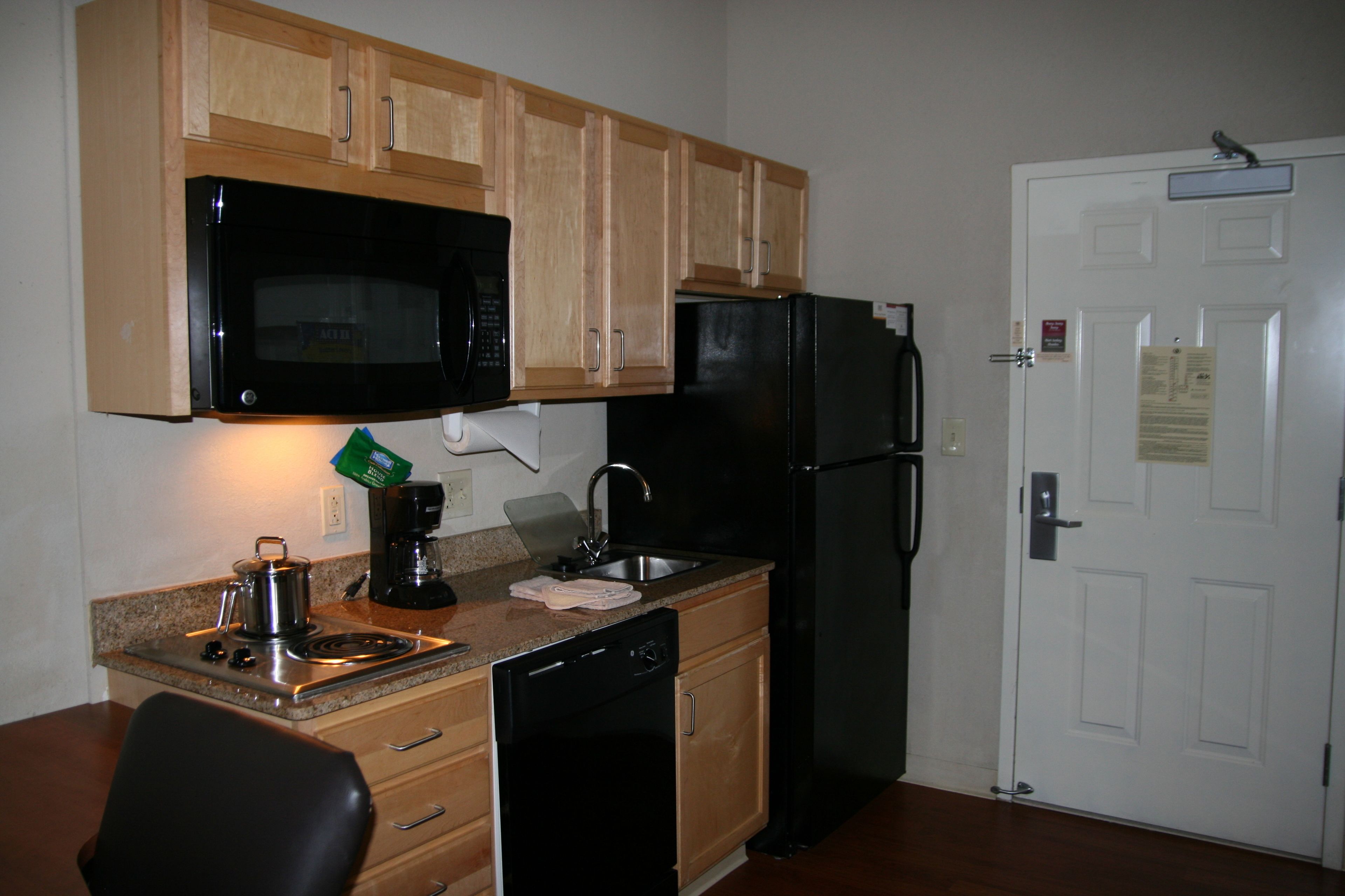 Cocina compacta con armarios de madera, microondas negro, refrigerador negro, cafetera y lavavajillas.