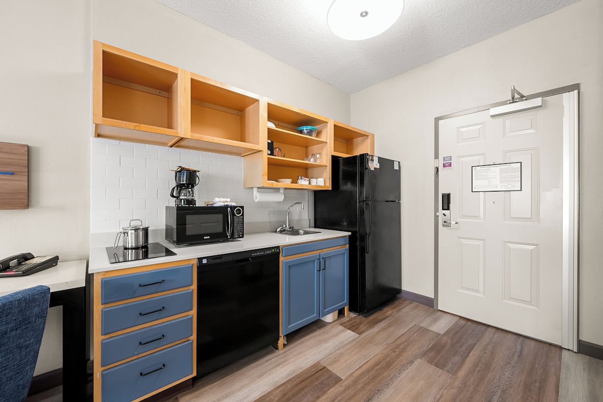 A compact kitchen with a microwave, coffee maker, and black refrigerator. There are blue cabinets and a wooden shelf above the sink.