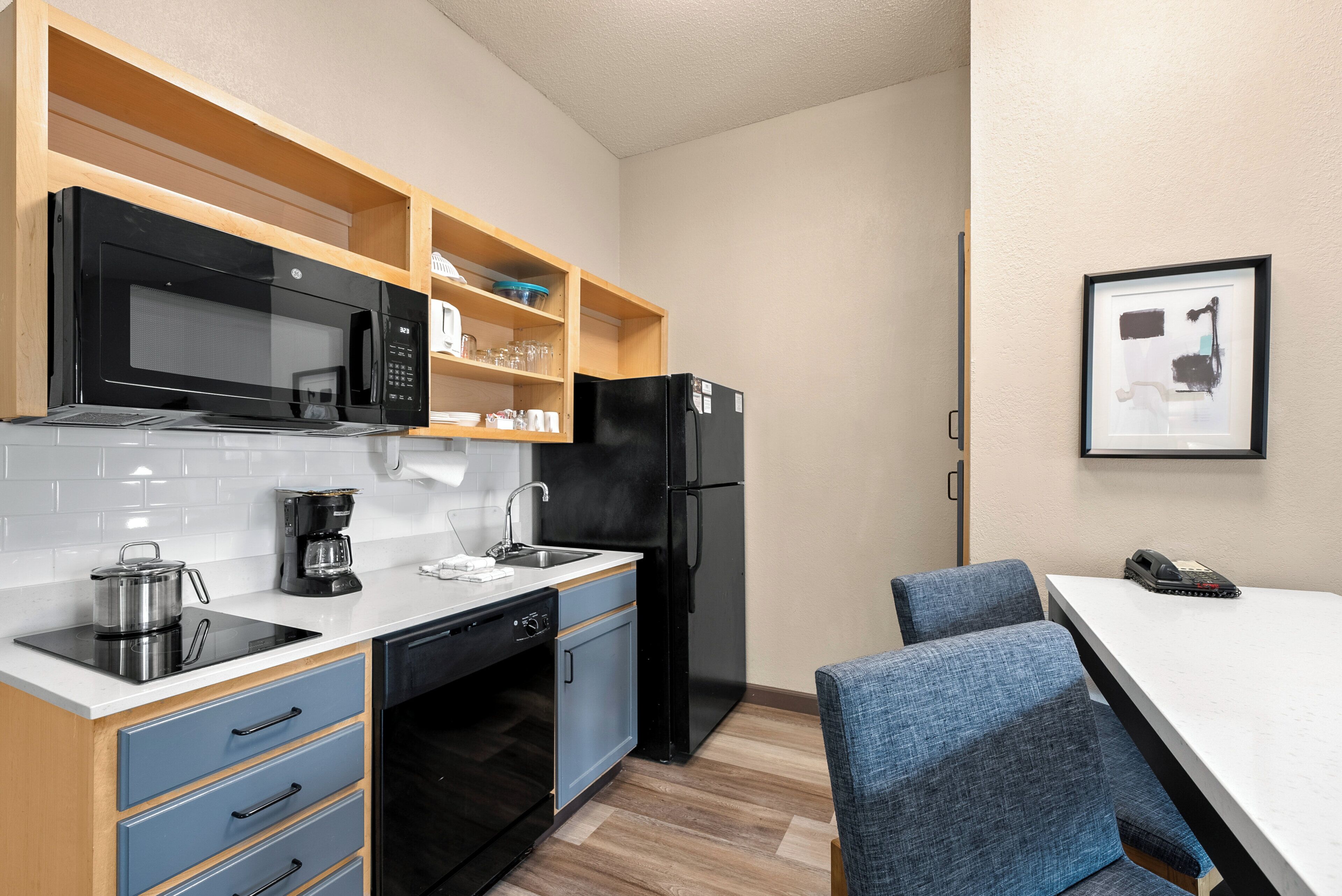 Una cocina compacta equipada con microondas, cafetera y refrigerador. Hay sillas azules y una pequeña mesa.