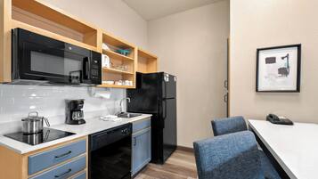 Una cocina compacta equipada con microondas, cafetera y refrigerador. Hay sillas azules y una pequeña mesa.