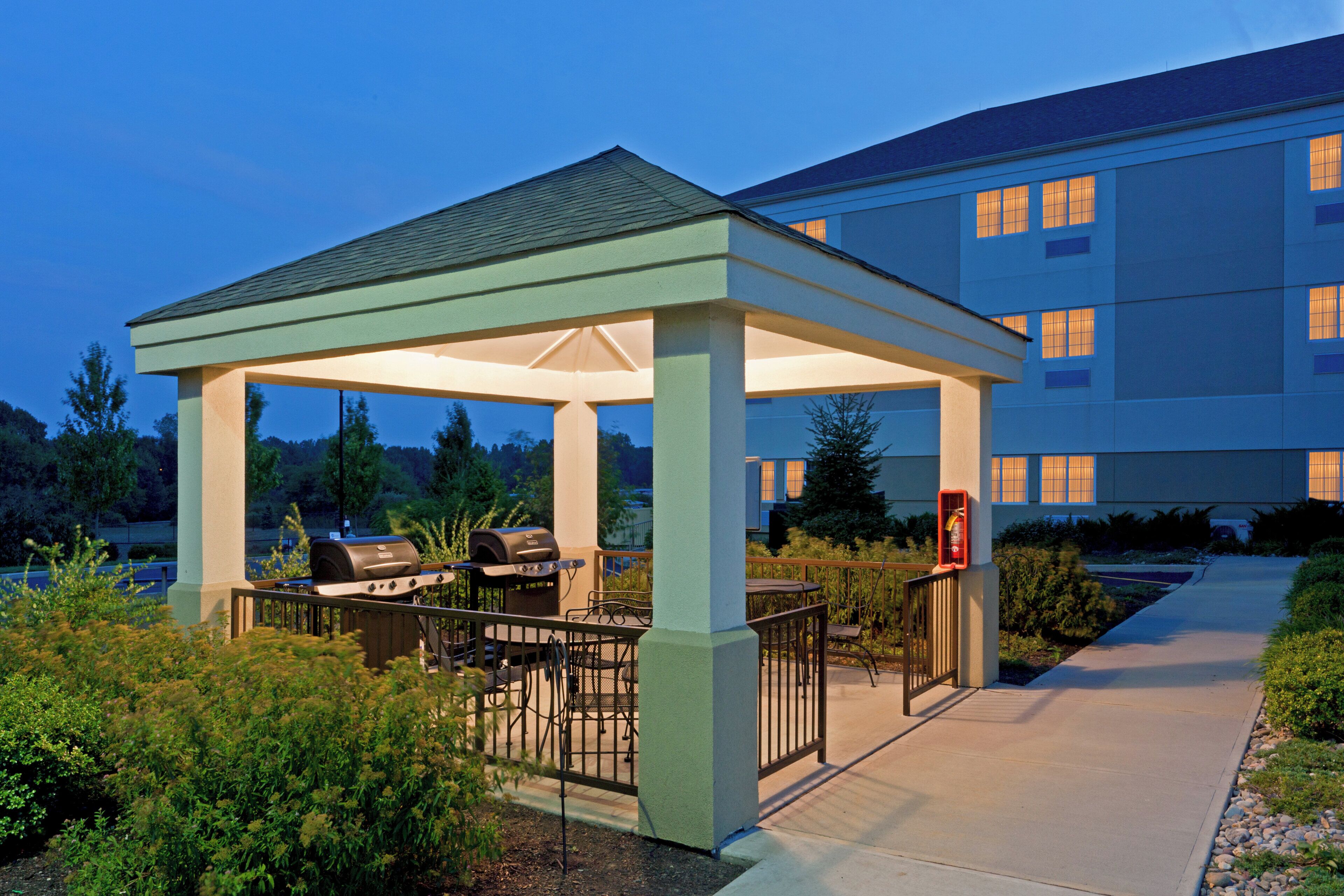 A covered outdoor seating area with barbecue equipment, adjacent to a building with multiple windows.