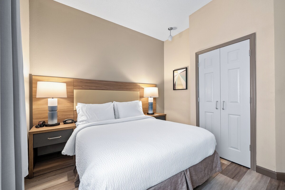 A neatly made bed with white bedding, two bedside lamps, a wooden headboard, a nightstand with a phone and a small table, and a white door.