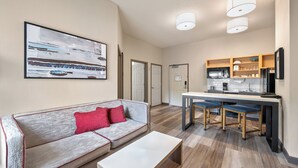 A modern living room with a grey sofa, red cushions, and a white coffee table. A kitchen area with a dining table and bar stools is visible in the background. - Candlewood Suites Bordentown-Trenton by IHG (Bordentown)
