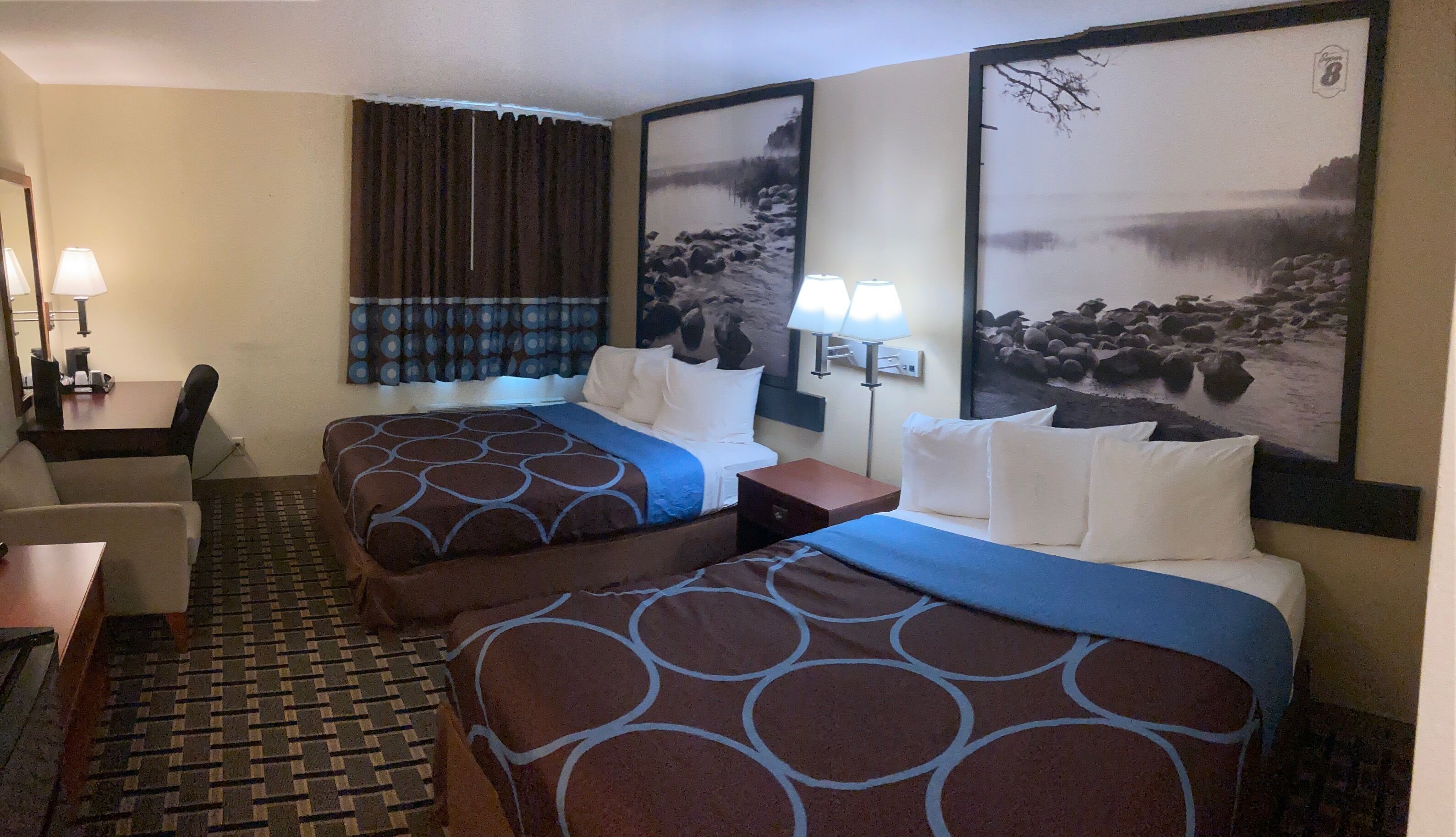 A hotel room with two beds, a desk, a chair, and two wall-mounted lamps.