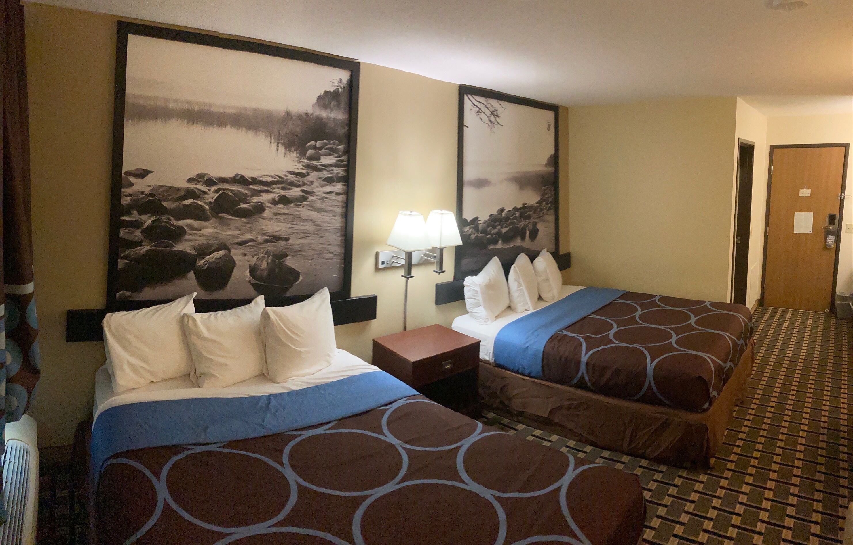 A hotel room with two beds, a nightstand, a lamp, and a door.