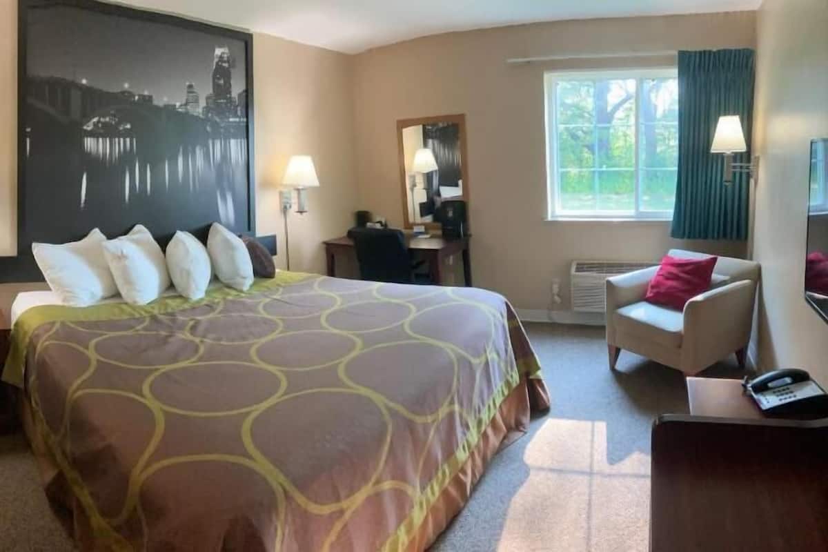 Hotellihuone, jossa on suuri sänky, kaksi yöpöytälamppua, työpöytä tuolilla, ikkuna verhoilla ja kaupunkimaisemaa esittävä taulu seinällä.