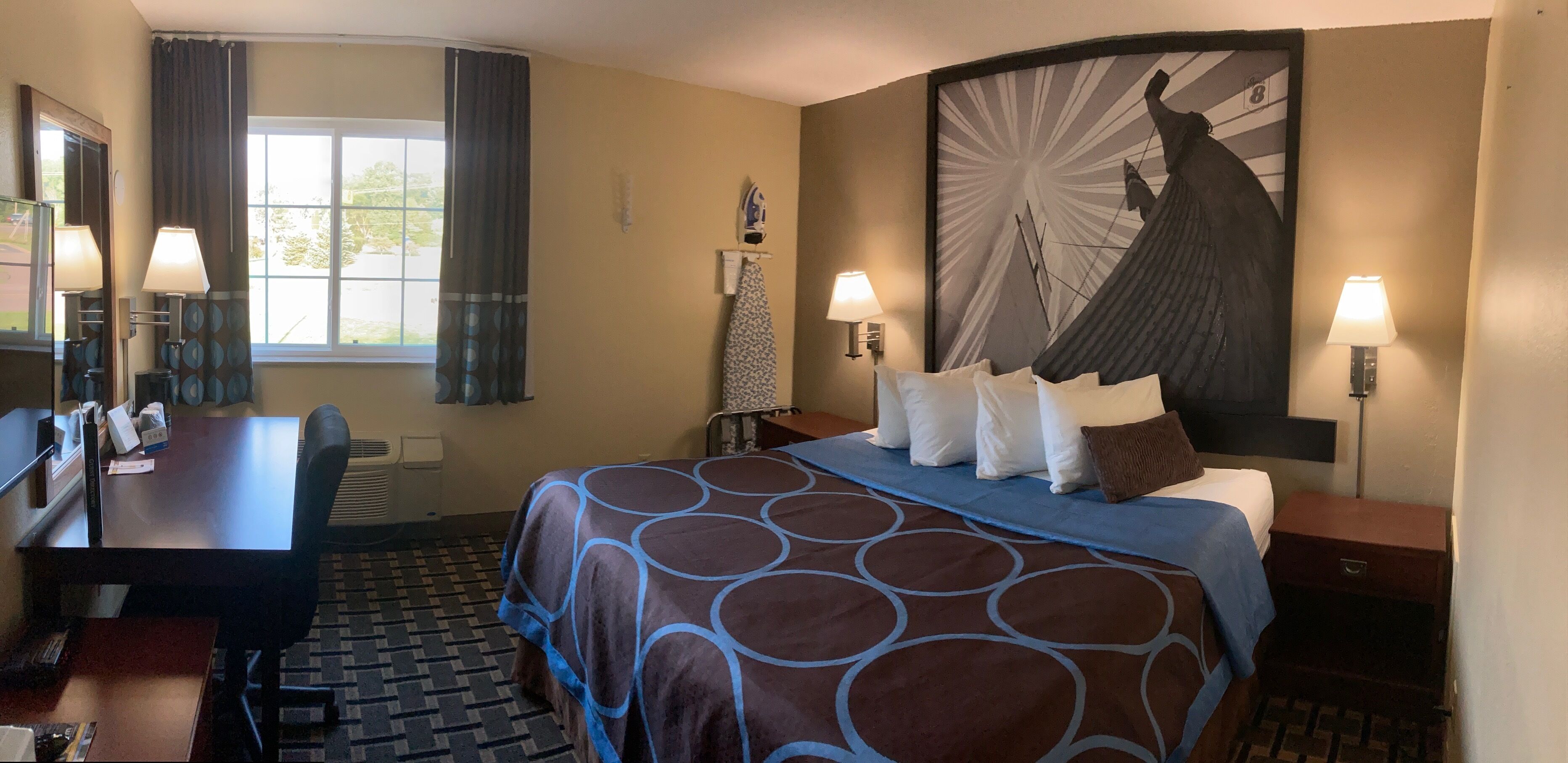 Une chambre d’hôtel avec un grand lit, un bureau et une chaise. Il y a deux lampes de chaque côté du lit et un grand tableau au mur.