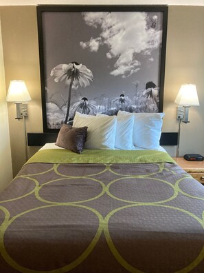 A hotel room with a bed, two bedside lamps, a large framed picture on the wall, and a digital clock on the nightstand.