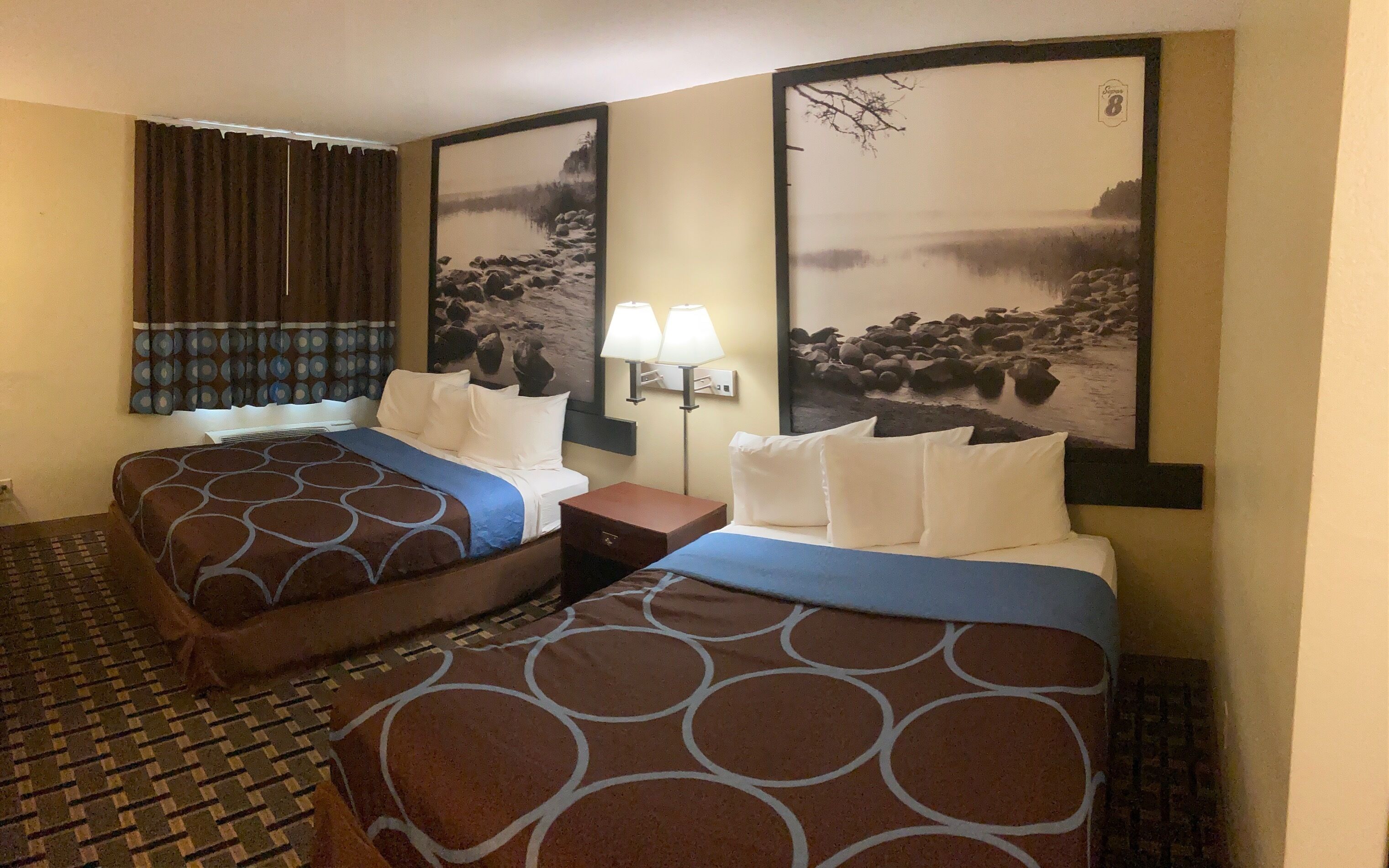 Une chambre d’hôtel avec deux lits, une table de chevet, une lampe et deux grandes peintures représentant des paysages accrochées aux murs.