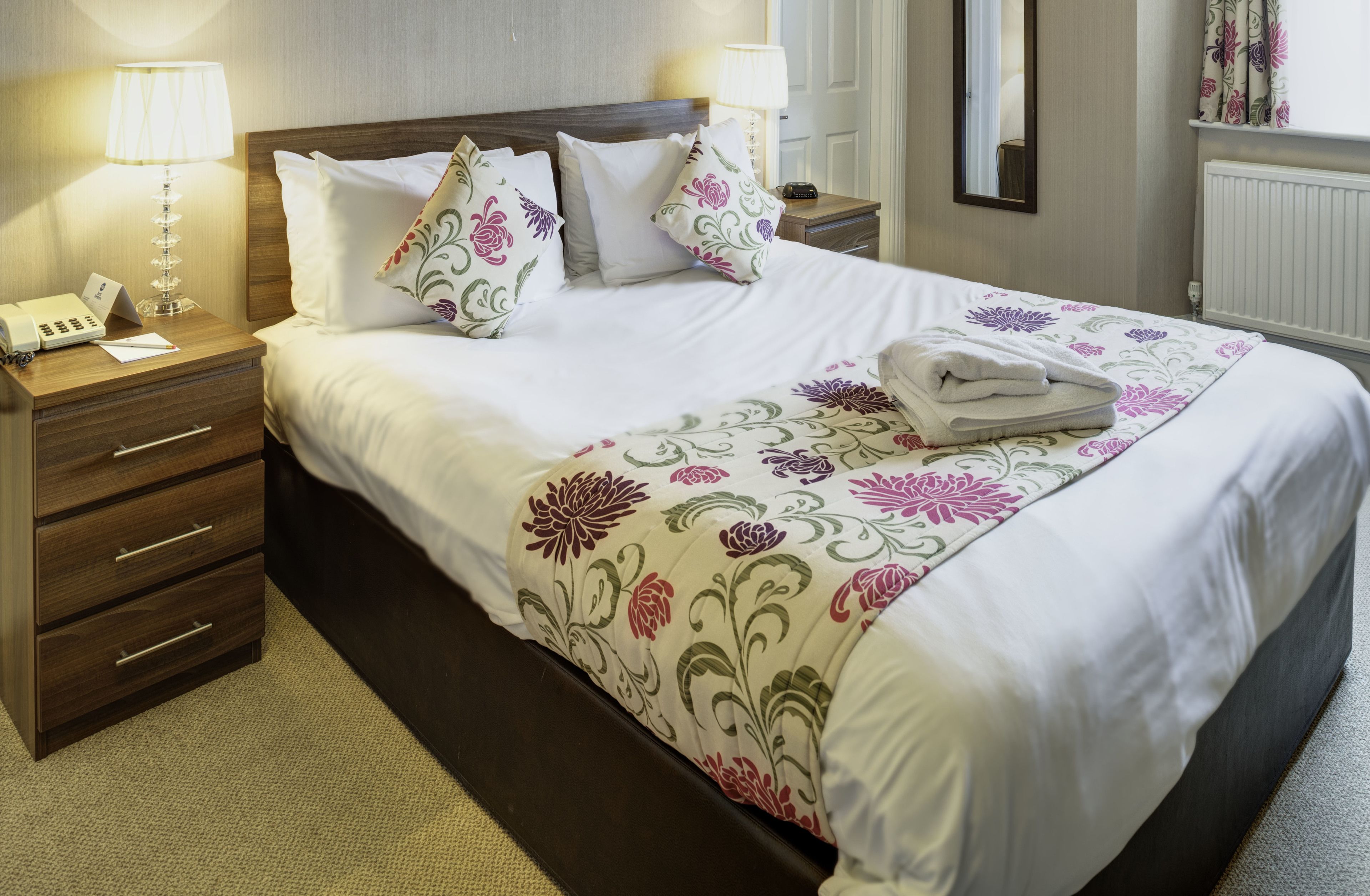 A neatly made bed with floral bedding, a bedside lamp, a telephone, and a folded towel on the nightstand.