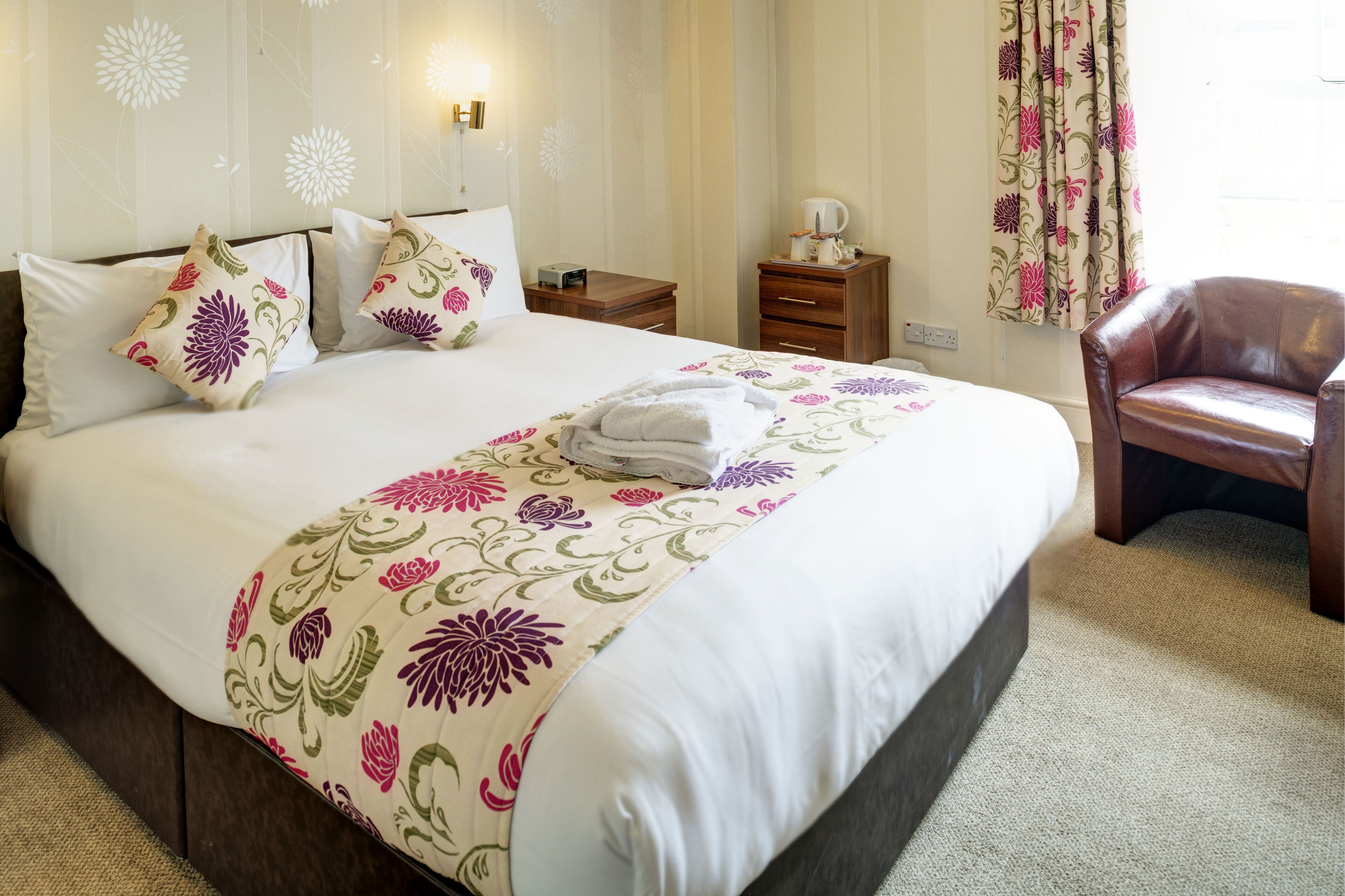 A neatly made bed with floral bedding, a wooden nightstand with a lamp, and a leather chair.