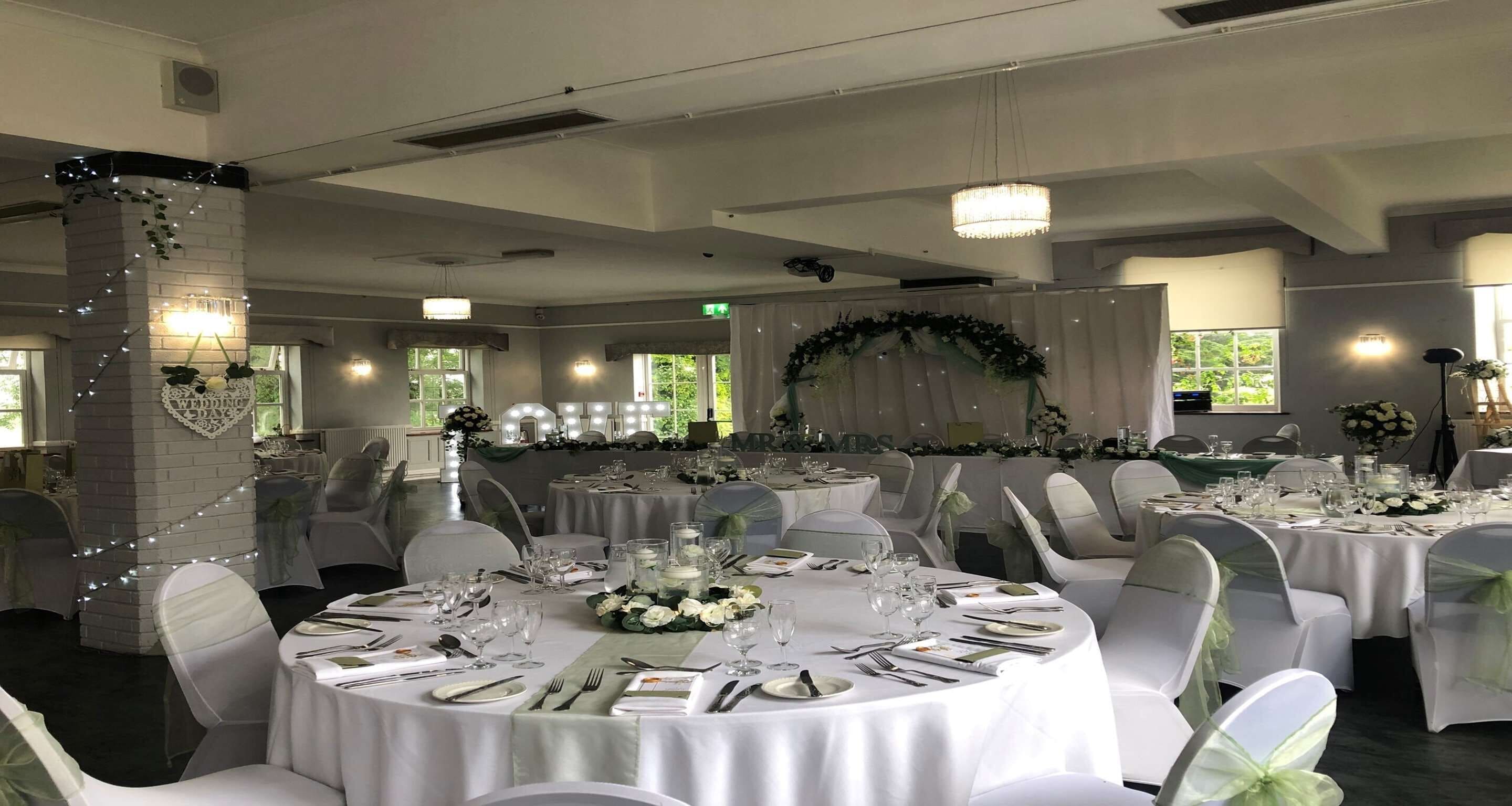 A banquet hall with round tables set for a formal event, white chairs, and a decorative arch with greenery.