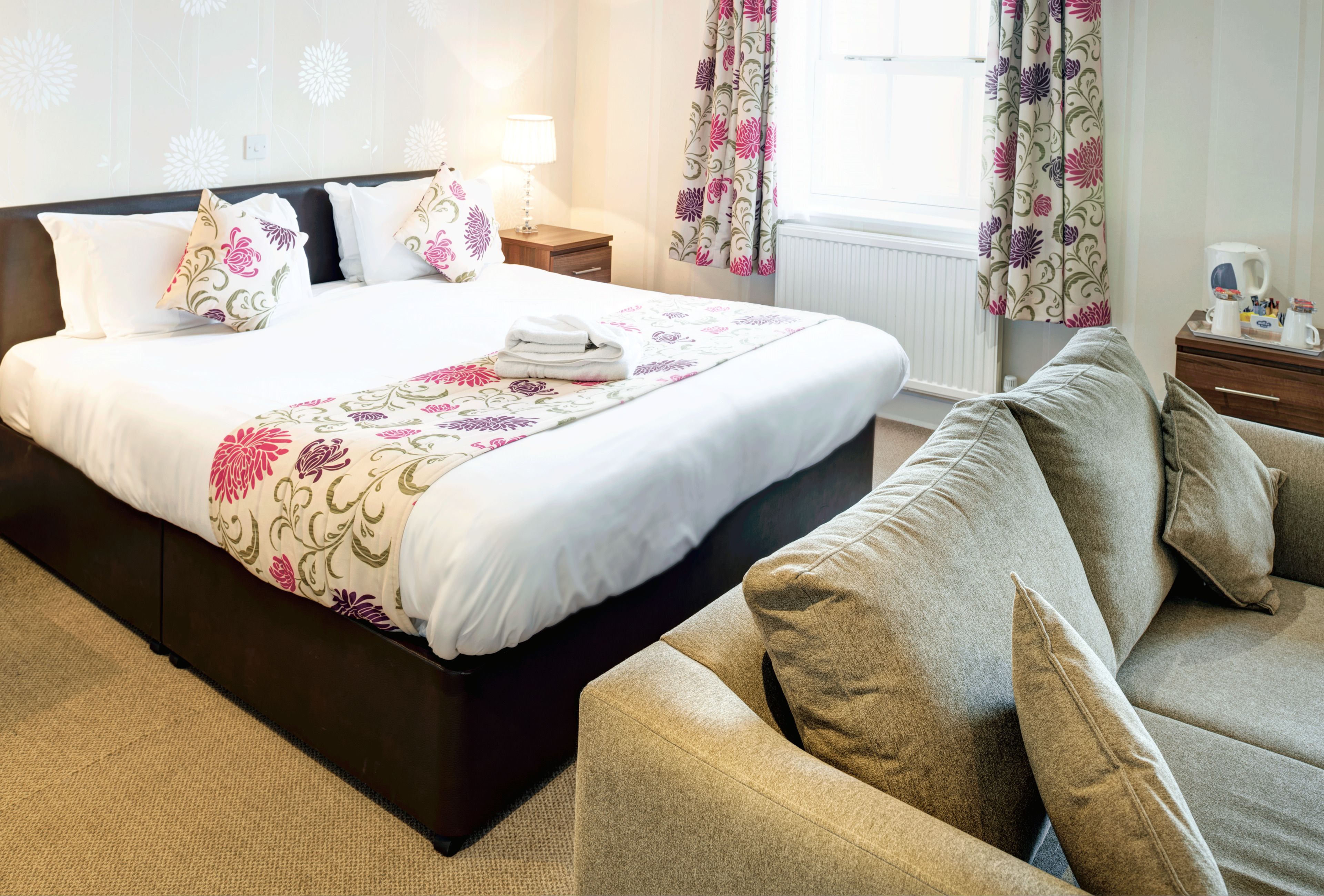 A neatly made bed with floral-patterned pillows and a blanket, a grey sofa with cushions, a bedside table with a lamp, and a window with floral curtains.