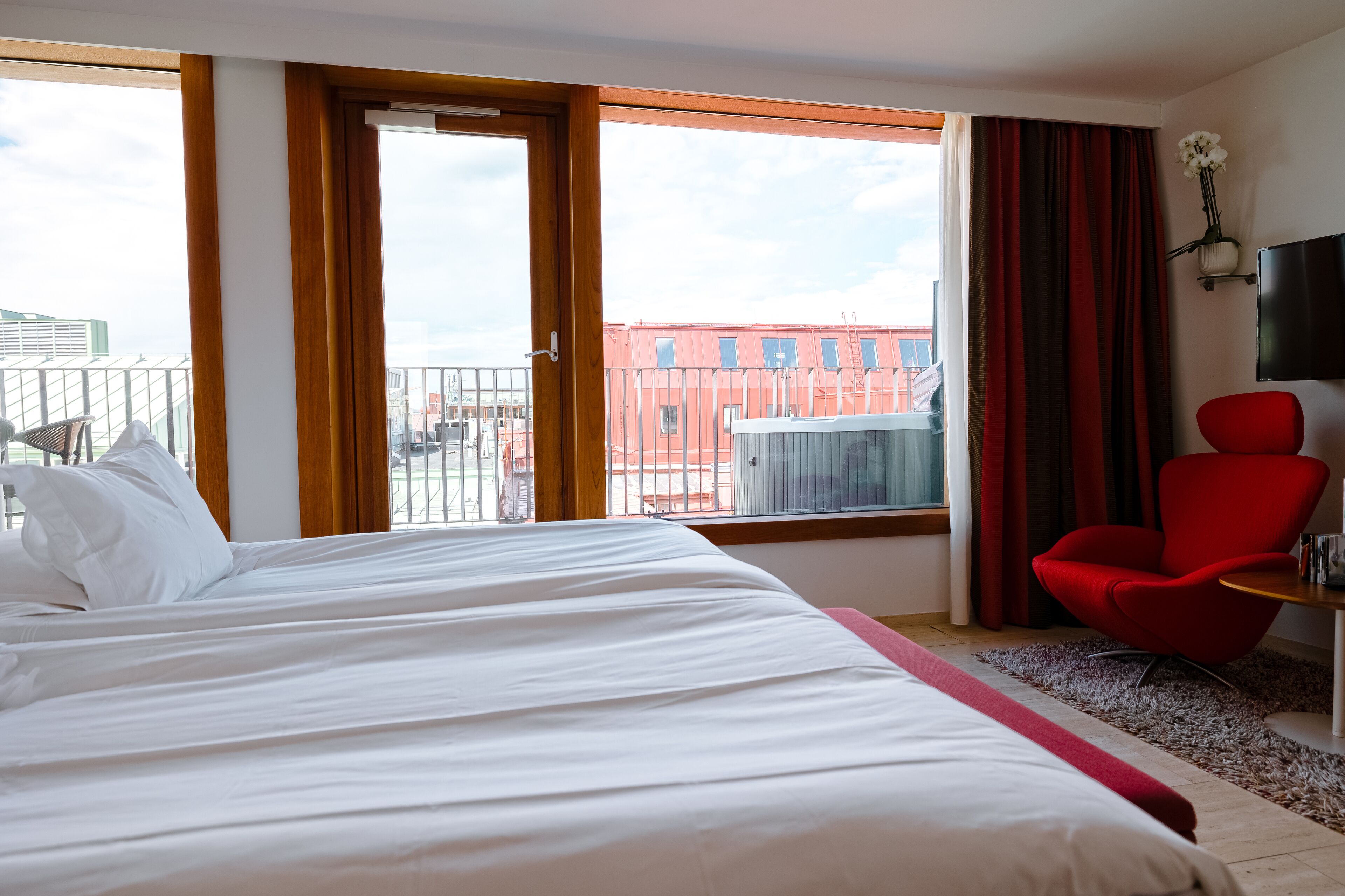 Una habitación de hotel con una cama grande, una silla roja, un televisor y vistas a edificios a través de la ventana.