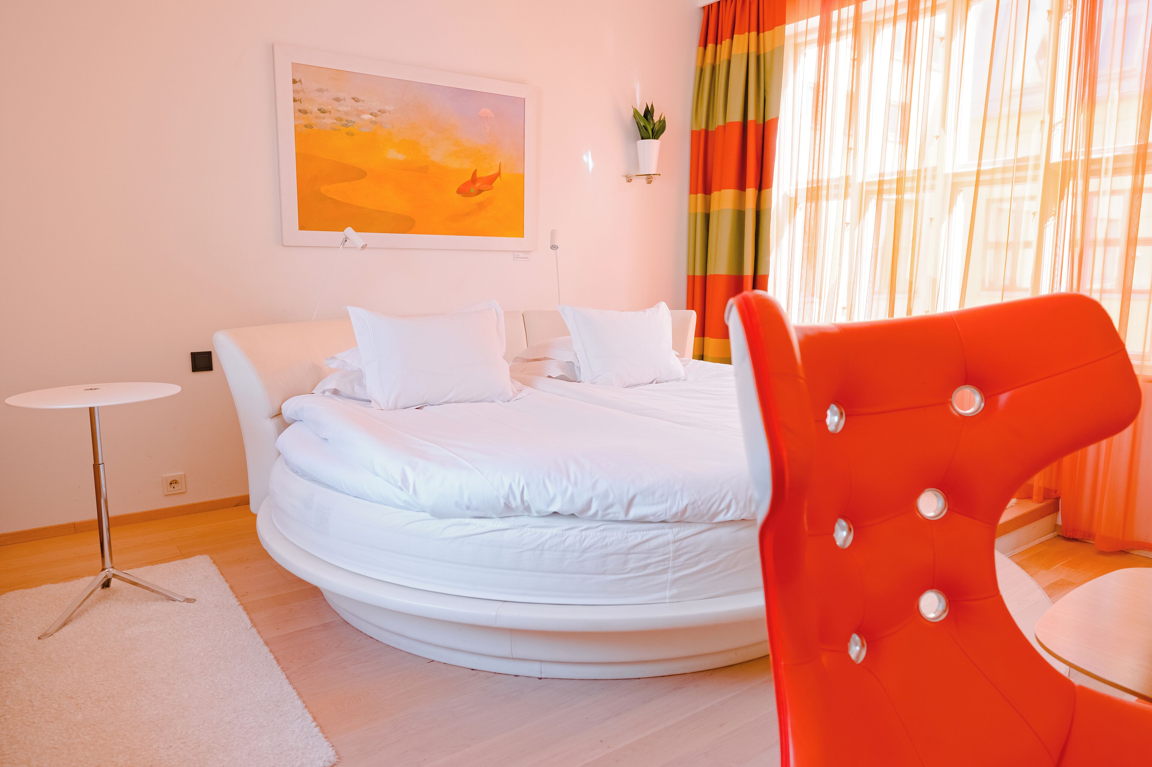 Un dormitorio con cama, una silla, una mesita, un cuadro y cortinas.