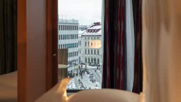 Ett hotellrum med en säng, ett fönster med gardiner och utsikt över byggnader utanför.