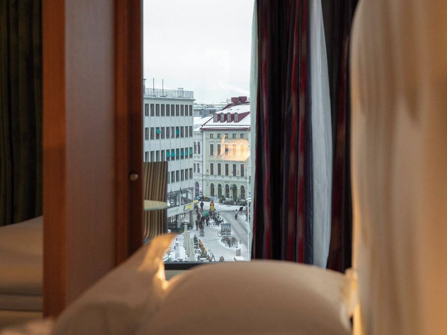 Ett hotellrum med en säng, ett fönster med gardiner och utsikt över byggnader utanför.