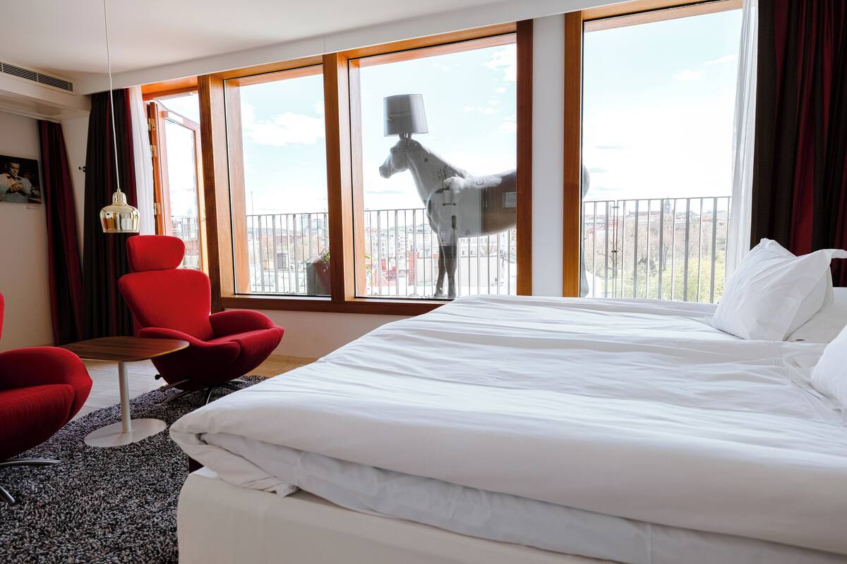 Ett hotellrum med en stor säng, en röd stol, ett litet bord och utsikt över en balkong med en häststaty.