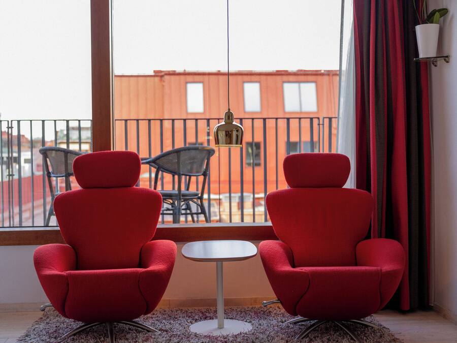 Varanda com duas cadeiras vermelhas e uma mesa pequena, com vista para um edifício vermelho.