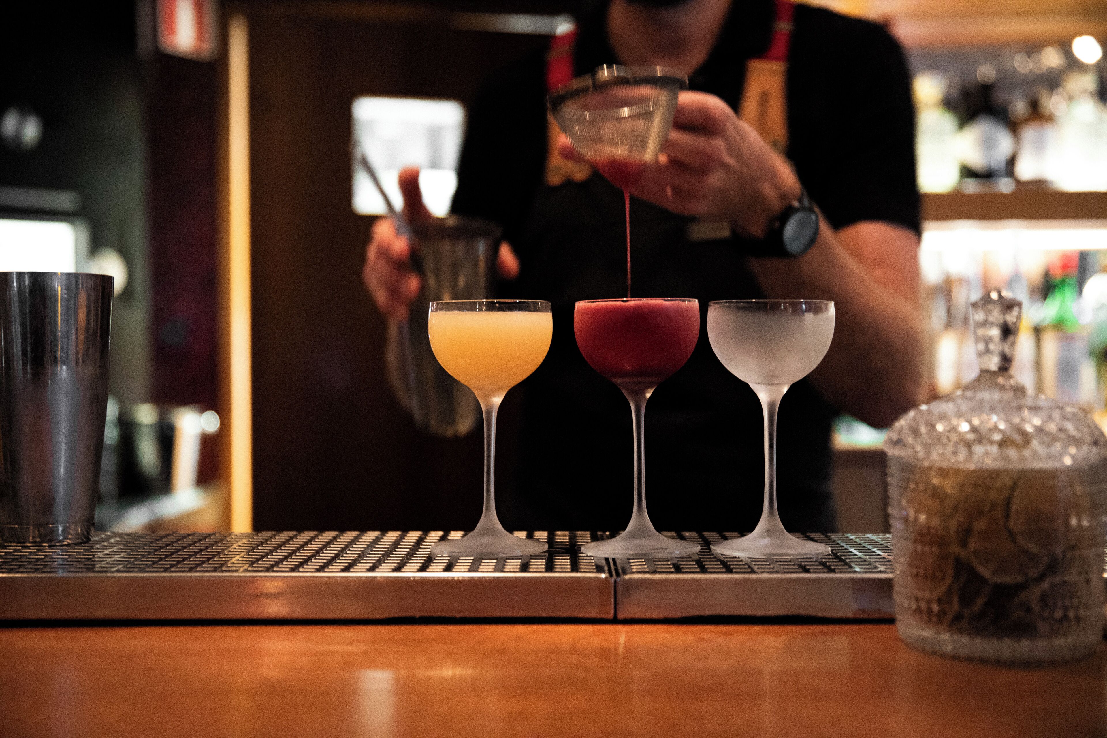 A bar area with three glasses of different colored drinks, a barista preparing a drink, and a coffee machine.
