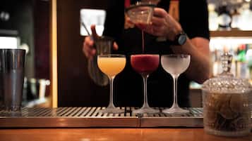 A bar area with three glasses of different colored drinks, a barista preparing a drink, and a coffee machine.