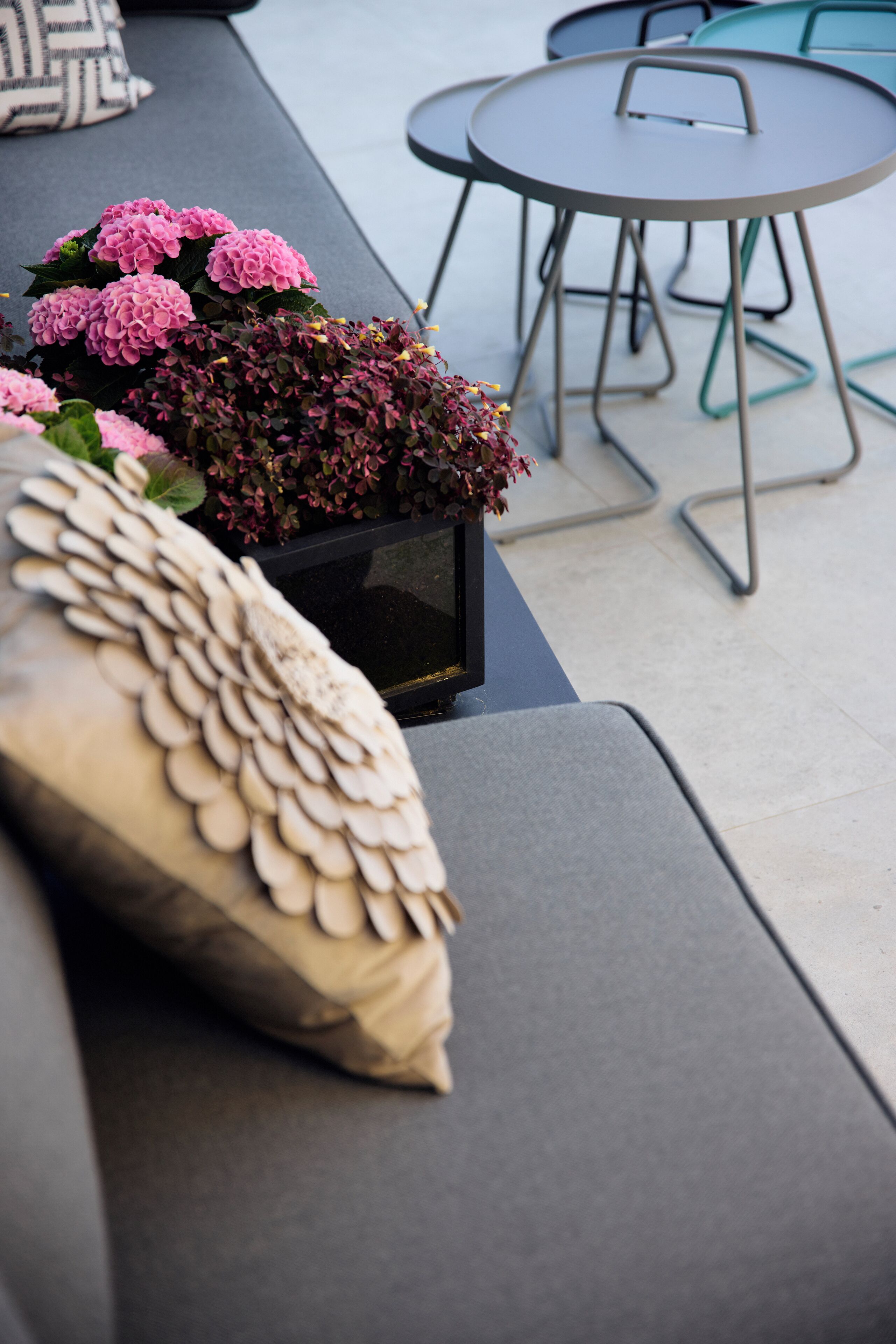 A modern outdoor seating area with a cushioned bench, a small table, and a potted plant with pink flowers.