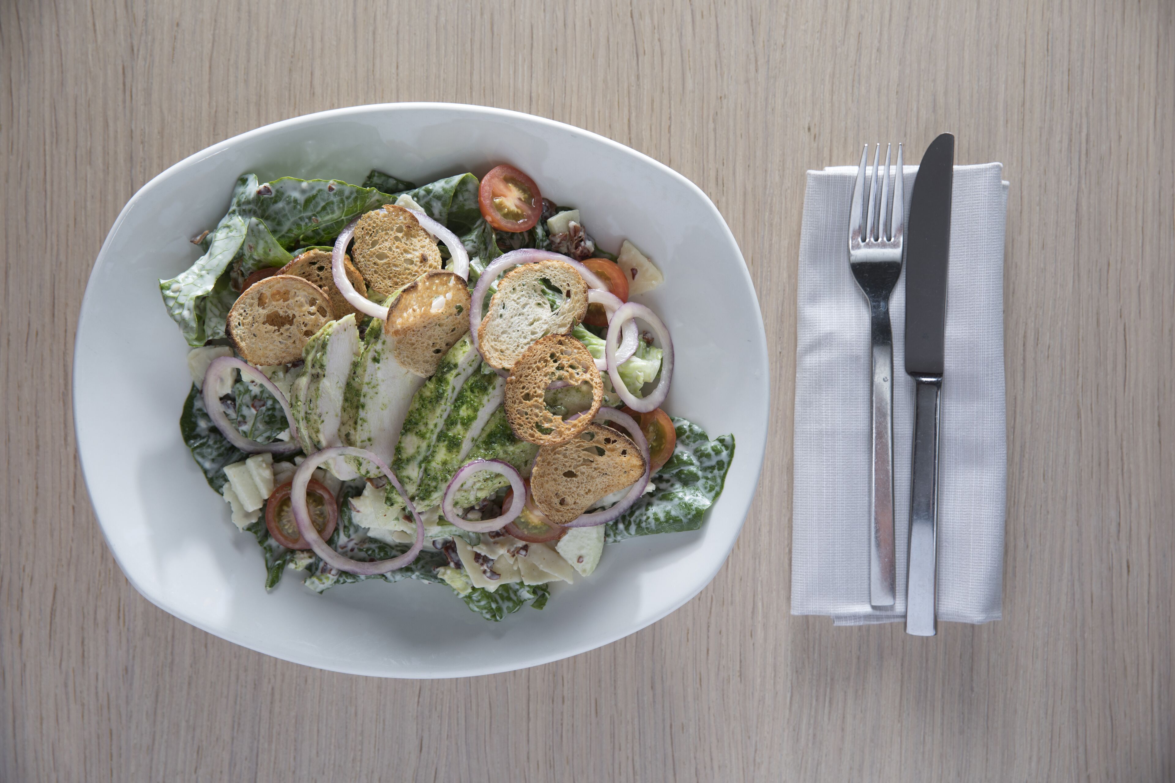 Un plato blanco ovalado con una ensalada, que incluye lechuga, tomates, cebollas y cubitos de pan, sobre una mesa de madera.