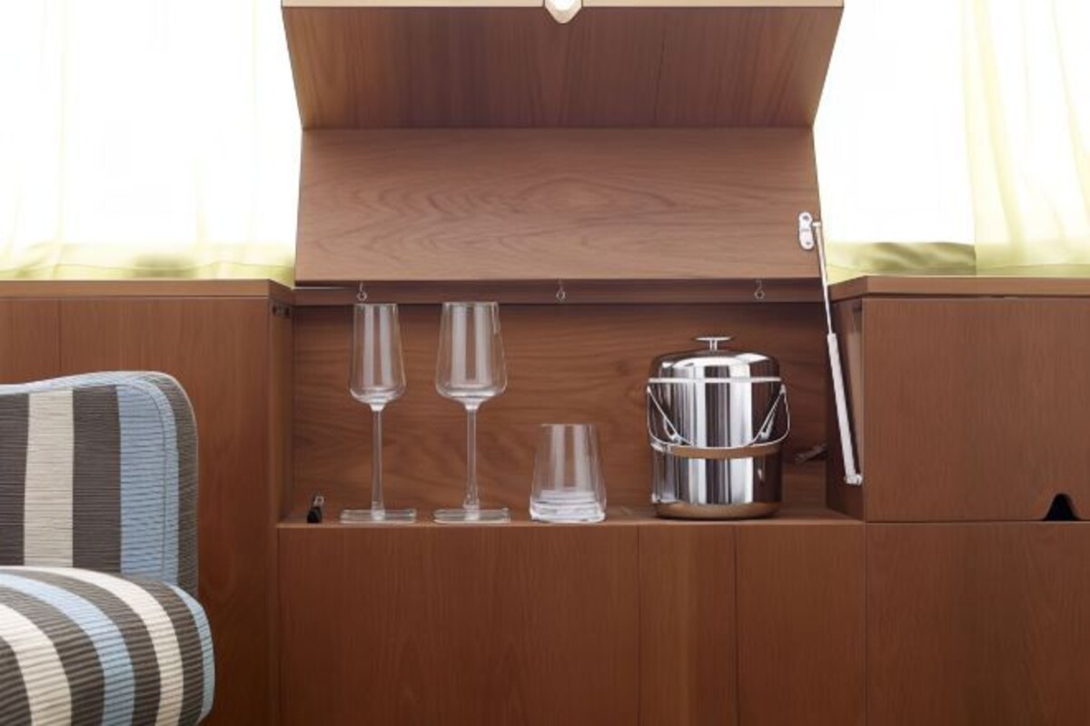 Un mueble de madera con tapa abatible que contiene dos copas de vino, un vaso pequeño y un recipiente de acero inoxidable.