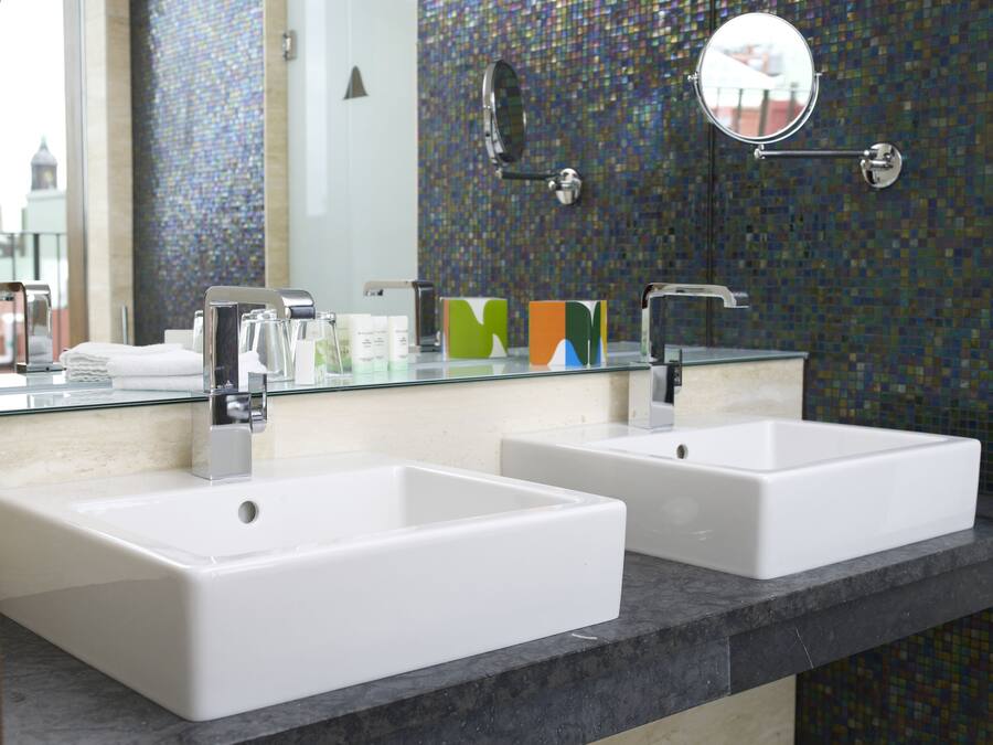 Une salle de bain avec deux lavabos blancs, un plan de toilette en verre et un miroir agrémenté d’un pare-feu en mosaïque.