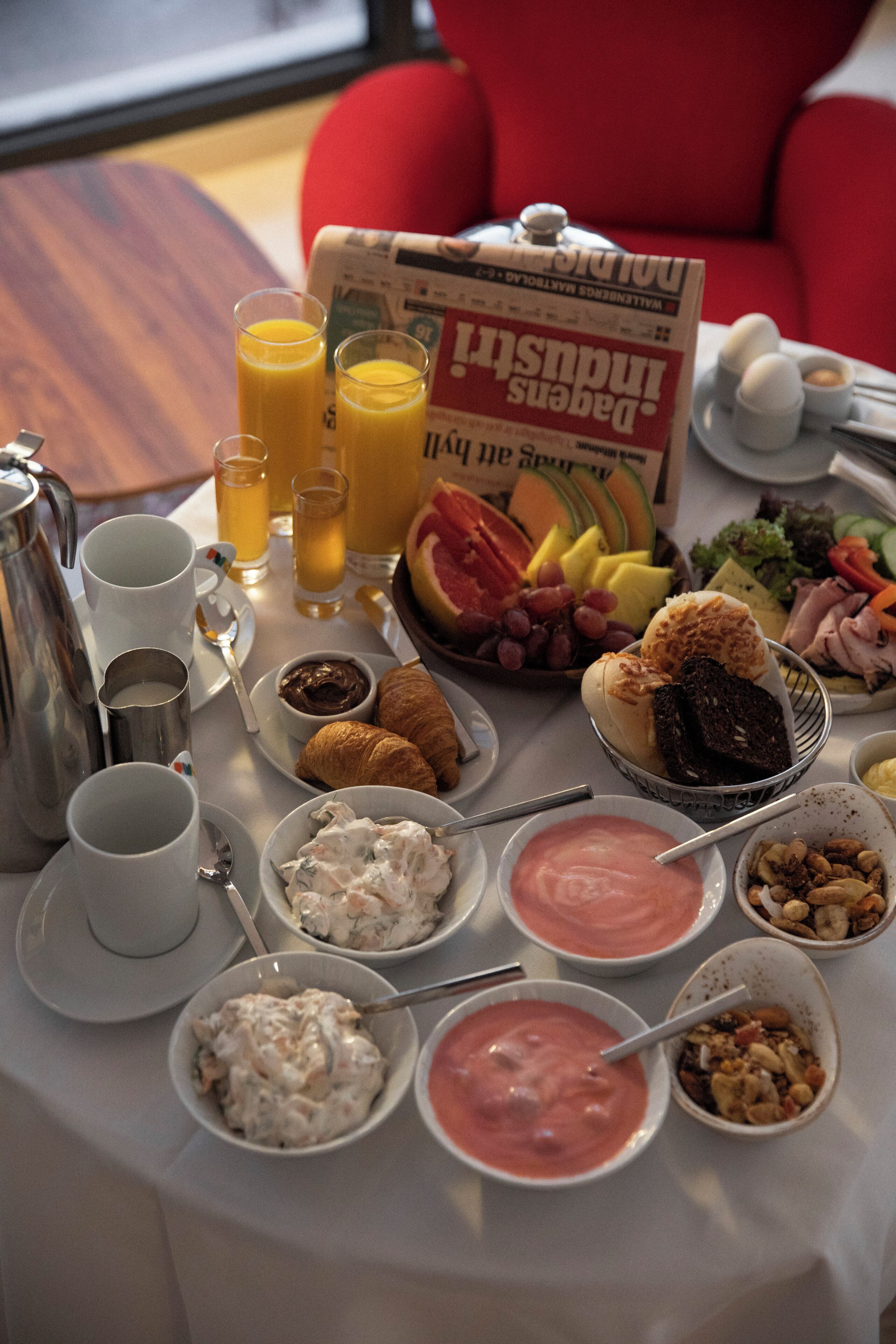 A breakfast table with various food items and drinks, including croissants, fruit, and a newspaper.
