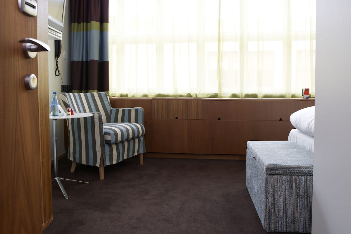 Ett hotellrum med en randig fåtölj, ett litet bord med en flaska och en telefon, en säng med ett grått täcke och ett fönster med tunna, genomskinliga gardiner.