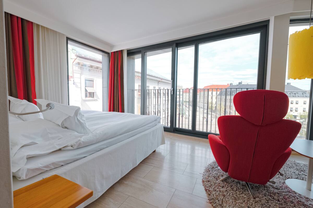 Ett modernt hotellrum med en stor säng, en röd fåtölj och utsikt över staden genom stora fönster.