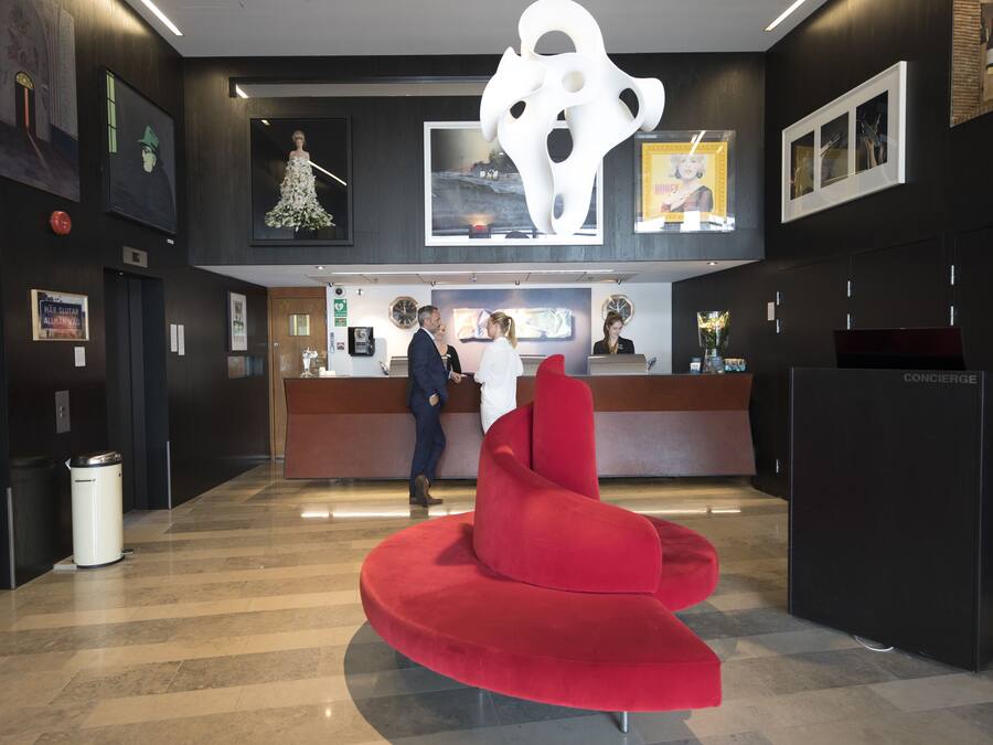 A hotel reception area with a modern design, featuring a red seating area, a concierge desk, and artwork on the walls.