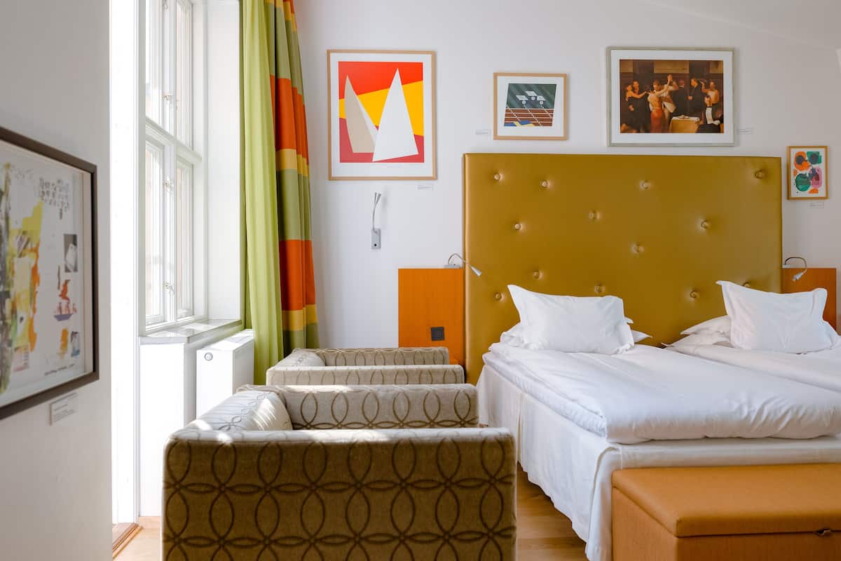 Ett hotellrum med en säng, två sittpallar, ett fönster med gardiner och konstverk på väggarna.