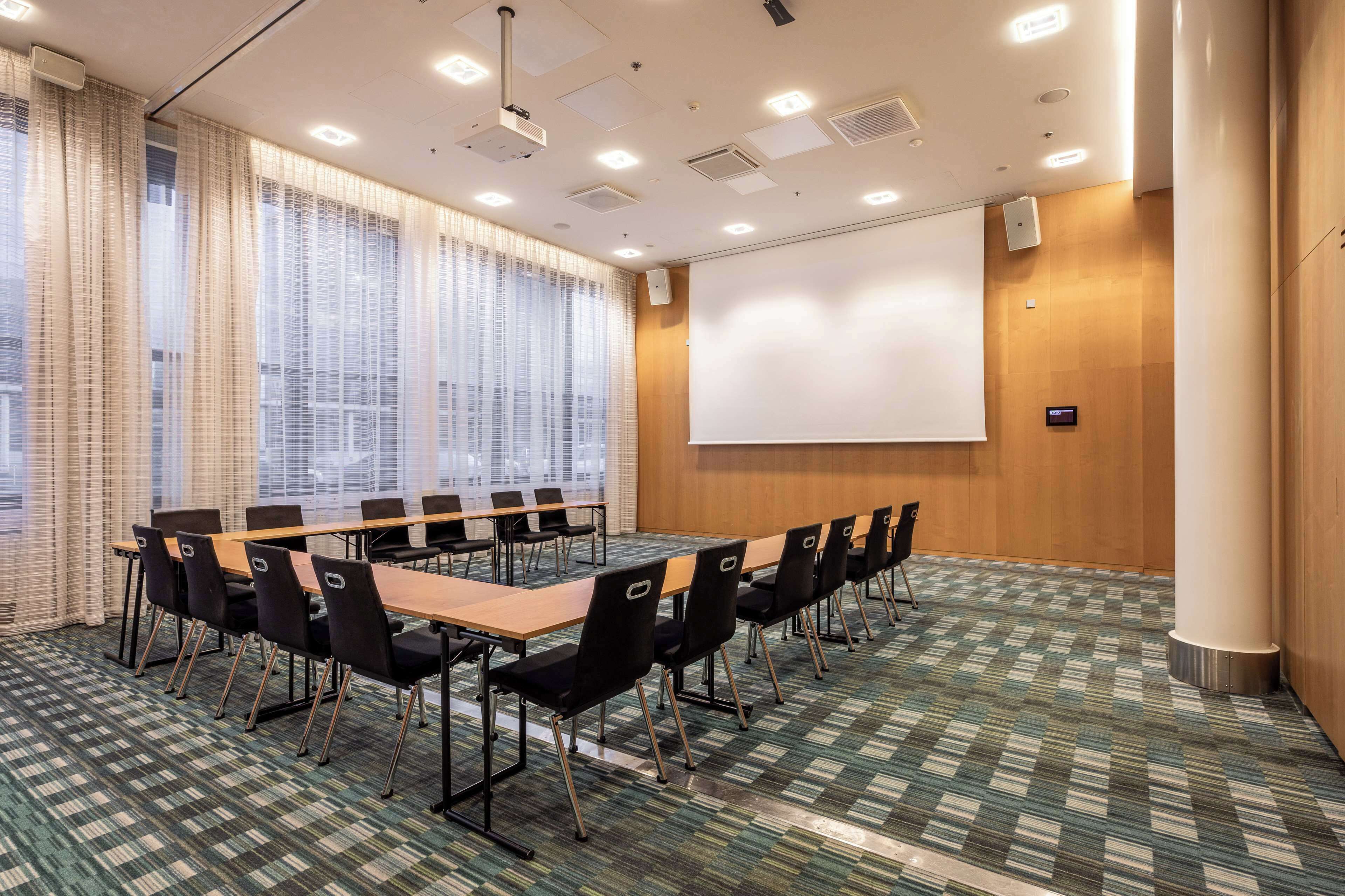 A conference room with a long table, chairs, a projector screen, and large windows with curtains.
